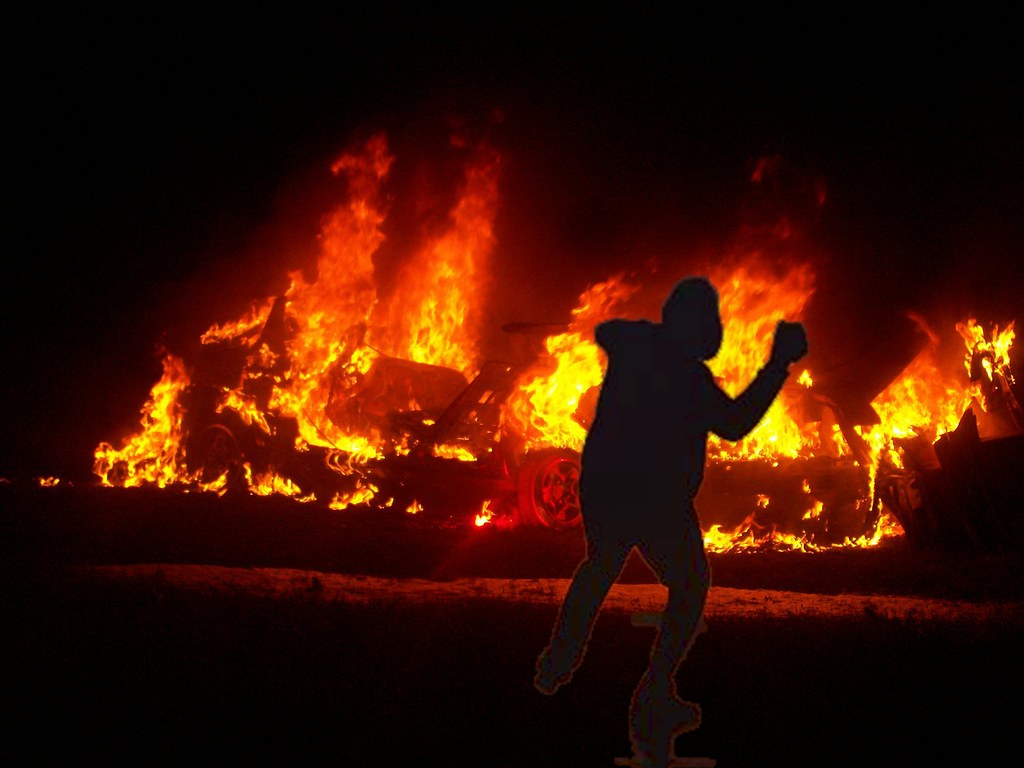 Silhouette einer werfenden Gestalt vor brennenden Autos.